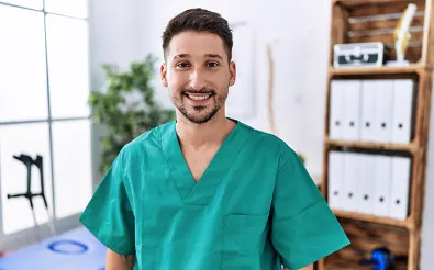 Ein Sporttherapeut in grüner Berufskleidung steht in einem Therapieraum; im Hintergrund Gehstützen, Wirbelsäulenmodell und Aktenordner.