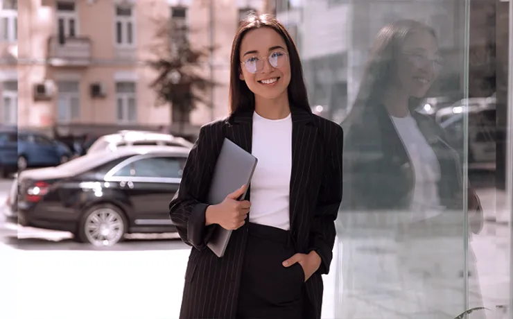 Eine Frau im dunklen Business-Outfit mit Brille steht lächelnd vor einem Glasgebäude und hält Unterlagen und ein Gerät in der Hand.