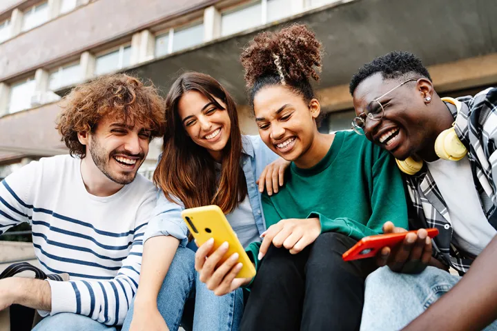Vier junge Menschen sitzen dicht nebeneinander auf einer Treppe vor einem Gebäude, schauen gemeinsam auf ein Smartphone und lachen.