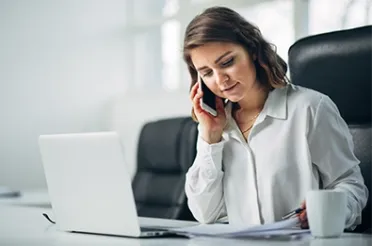 Eine Frau in weißer Bluse telefoniert an ihrem Schreibtisch und notiert dabei etwas; vor ihr stehen ein Laptop und eine Kaffeetasse.