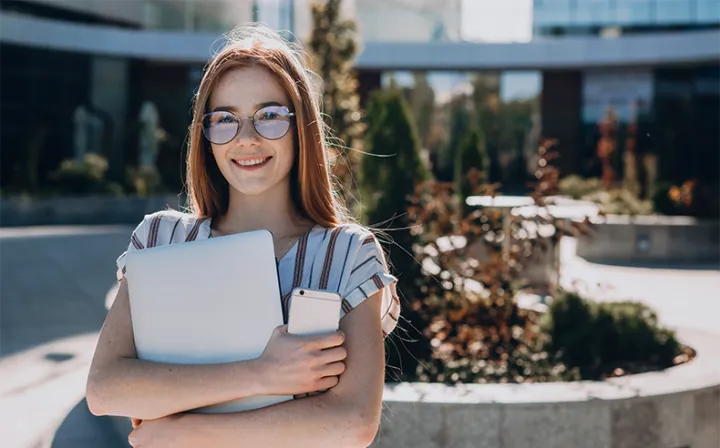 Eine junge Frau mit Brille steht mit Laptop und Smartphone im Arm vor einem Bürogebäude und schaut in die Kamera.