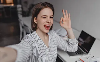 Eine junge Frau mit kurzem Haar zwinkert in die Kamera, zeigt das OK-Zeichen und macht ein Selfie an ihrem Schreibtisch mit Laptop.