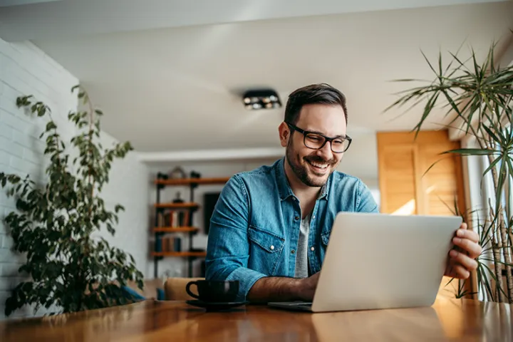 Ein Mann mit Brille sitzt lachend an einem Holztisch vor einem Laptop; neben ihm eine Kaffeetasse, im Raum mehrere Zimmerpflanzen.
