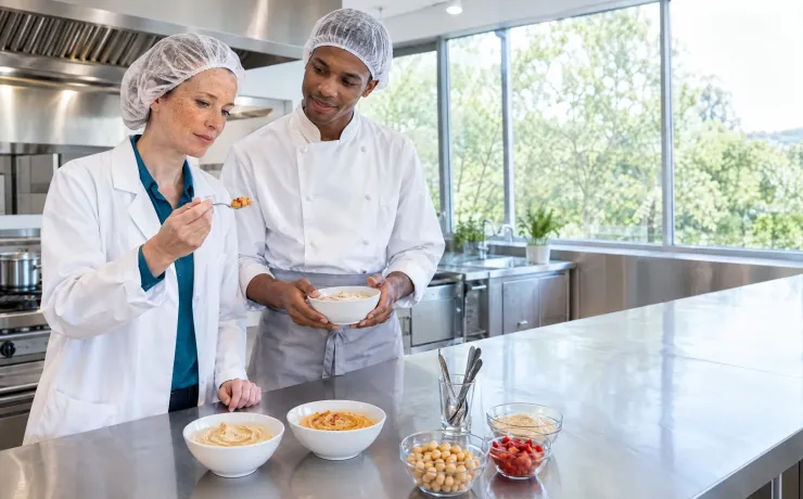 Produktmanagerin betrachtet in der Musterküche einen Probierlöffel mit der dunkleren Variante eines Kichererbsen-Aufstrichs, der Produktentwickler hält eine weitere Variante in der Schale