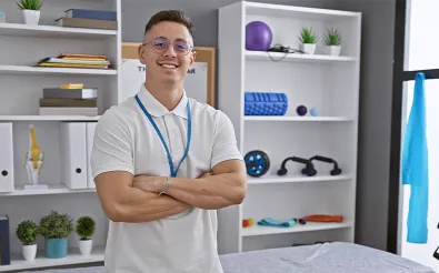 Ein Therapeut in weißem Poloshirt mit Namensband steht lächelnd mit verschränkten Armen in einem Praxisraum mit Regalen voller Trainingsgeräte.