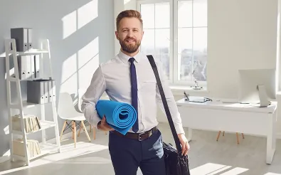 Ein Mann in Hemd und Krawatte mit Umhängetasche hält in einem Büro eine zusammengerollte blaue Gymnastikmatte.