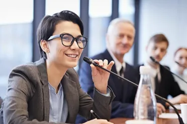 Eine Frau mit Brille spricht lächelnd in ein Mikrofon, hinter ihr sitzen weitere Personen an einem Tisch.
