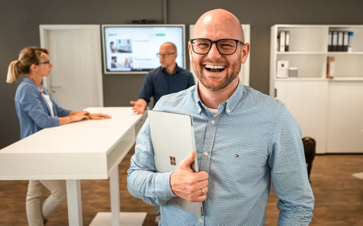 Ein Mann mit Brille lacht mit einem Laptop unter dem Arm in die Kamera; im Hintergrund zwei Kolleginnen und Kollegen an einem Stehtisch.