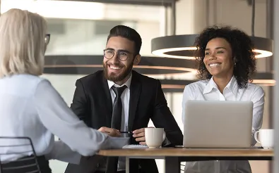 Ein Mann im Anzug und eine Frau in weißer Bluse sitzen lächelnd mit einer Gesprächspartnerin am Besprechungstisch, vor ihnen stehen ein Laptop und Kaffeetassen.