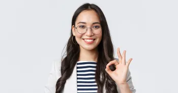 Eine junge Frau mit Brille lächelt in die Kamera und zeigt mit der Hand das Okay-Zeichen.