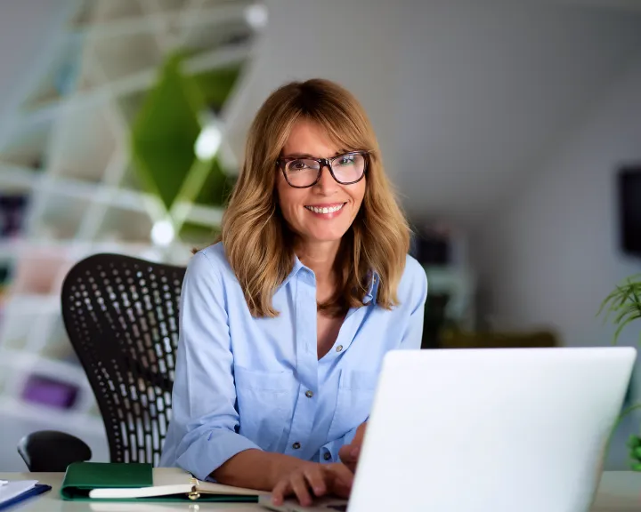 Eine Frau mit Brille sitzt am Schreibtisch vor einem Laptop und schaut lächelnd in die Kamera.