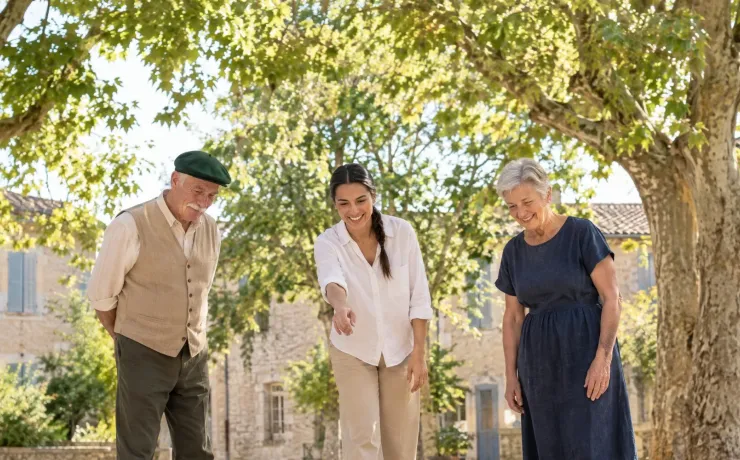 Eine Frau steht nach ihrem Wurf beim Boule auf einem Dorfplatz in der Provence, zwei ältere Nachbarn nicken anerkennend zu ihrer Kugel am Ziel