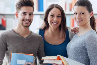 Drei Studierende schauen lachend in die Kamera & halten ihre Bücher in der Hand