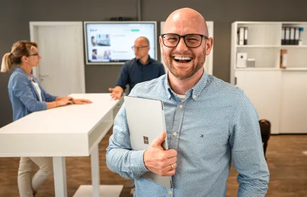 Ein Mann mit Brille lacht mit einem Laptop unter dem Arm in die Kamera; im Hintergrund zwei Kolleginnen und Kollegen an einem Stehtisch.
