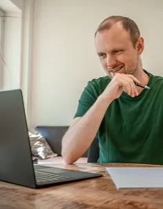 Ein lächelnder Mann sitzt mit Kugelschreiber in der Hand vor einem Laptop an einem Holztisch.