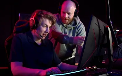 Ein junger Mann spielt mit Headset am Rechner, ein zweiter Mann zeigt ihm etwas auf dem Bildschirm.