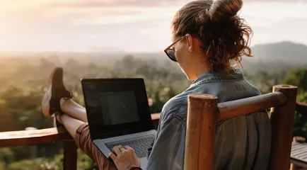 Eine Frau sitzt mit hochgelegten Beinen auf einer Terrasse über einer Berglandschaft und arbeitet am Laptop.