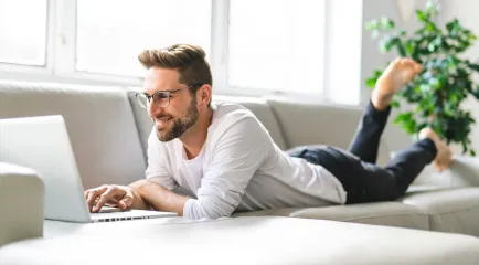 Ein lächelnder Mann mit Brille liegt auf einem Sofa und arbeitet an einem Laptop.