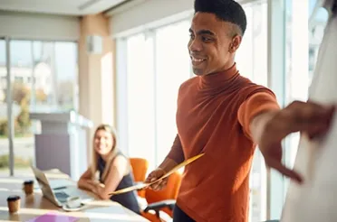 Ein Mann im orangefarbenen Rollkragenpullover geht lächelnd durch ein helles Büro und zeigt mit einer Mappe in der Hand die Richtung; im Hintergrund sitzt eine Kollegin am Laptop.