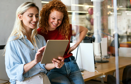 Zwei Frauen stehen nebeneinander in einem Büro und schauen gemeinsam lächelnd auf ein Tablet.