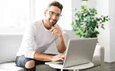 Ein Mann mit Brille sitzt lächelnd auf einem Sofa und arbeitet am Laptop.
