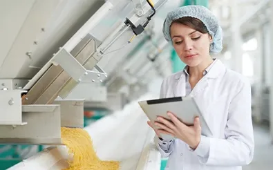 Eine Frau mit Haarnetz und weißem Kittel prüft in einer Produktionsanlage Daten auf einem Tablet, neben ihr läuft ein Förderband.