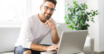 Ein Mann mit Brille sitzt entspannt auf einem hellen Sofa, lächelt in die Kamera und arbeitet am Laptop; hinter ihm steht eine Zimmerpflanze.