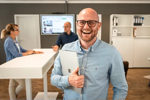 Ein lachender Mann hält ein Tablet unter dem Arm, hinter ihm sprechen zwei Personen in einem Büro.