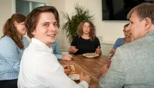 Mehrere Menschen sitzen bei einem gemeinsamen Essen an einem Holztisch, im Vordergrund lächelt ein junger Mann.