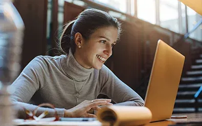 Eine junge Frau lächelt beim Lernen an einem Laptop, neben ihr liegen Papierrollen und Stifte auf dem Tisch.