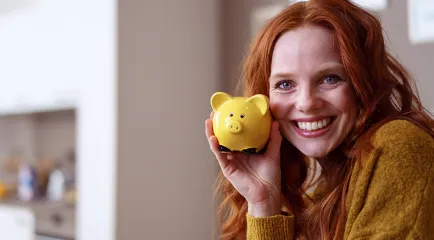 Eine lächelnde Frau hält ein gelbes Sparschwein an ihre Wange und steht für die Finanzierung des Studiums.