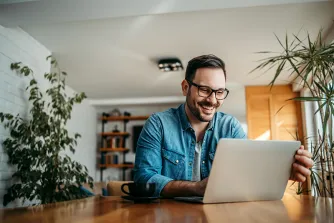Ein Mann mit Brille sitzt lachend an einem Holztisch vor einem Laptop; neben ihm eine Kaffeetasse, im Raum mehrere Zimmerpflanzen.
