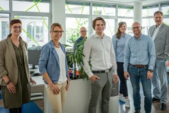 Ein Team aus sieben Mitarbeitenden steht lächelnd in einem hellen Büro mit Pflanzen und Monitoren.