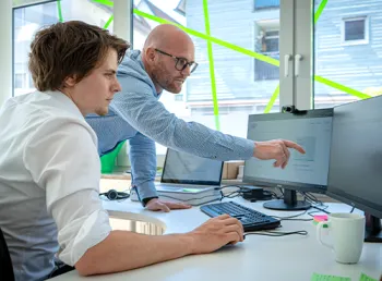 Ein Mann zeigt einem jungen Kollegen etwas auf dem Monitor an einem gemeinsamen Arbeitsplatz.