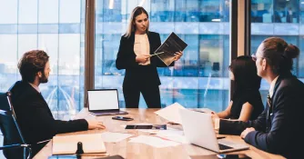 Eine Frau in Business-Kleidung präsentiert in einem Besprechungsraum mit Glasfront Unterlagen vor drei sitzenden Kollegen.
