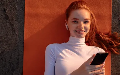 Eine junge Frau mit Kopfhörern lehnt an einer orangefarbenen Wand und hält ein Smartphone in der Hand.
