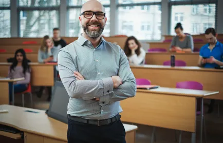 Ein Dozent mit Brille steht lächelnd mit verschränkten Armen in einem Hörsaal, an den Tischen dahinter sitzen Studierende.