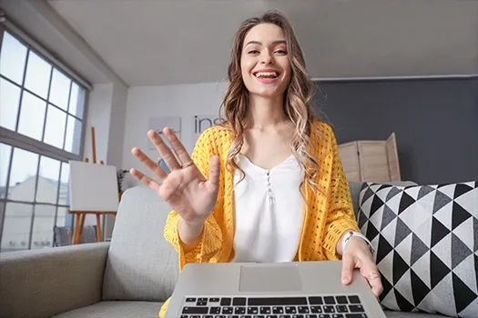 Eine Frau sitzt vor einem Laptop auf dem Sofa und winkt lachend in die Kamera.