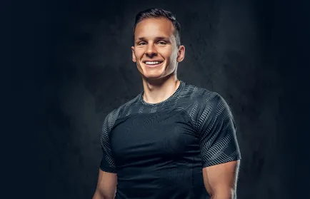 Ein lächelnder, muskulöser Mann im dunklen Sportshirt steht vor dunklem Hintergrund und blickt in die Kamera.