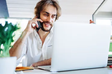 Ein Mann mit Bart telefoniert lächelnd vor seinem Laptop an einem hellen Arbeitsplatz mit Zimmerpflanzen.