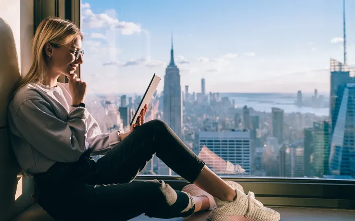 Eine junge Frau sitzt am Fenster eines Hochhauses mit Blick auf eine Großstadt und liest auf einem Tablet.