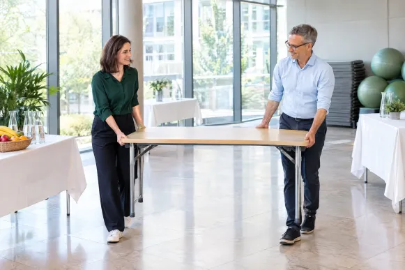 Organisatorin und Betriebsleiter tragen im Foyer beim Aufbau eines Gesundheitstags gemeinsam einen Klapptisch zwischen die Stationen