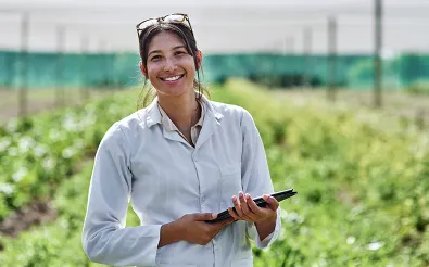 Eine lächelnde Frau im weißen Kittel steht mit einem Tablet auf einem Feld mit Nutzpflanzen.