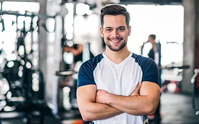 Trainer steht lächelnd mit verschränkten Armen zwischen Geräten im Fitnessstudio