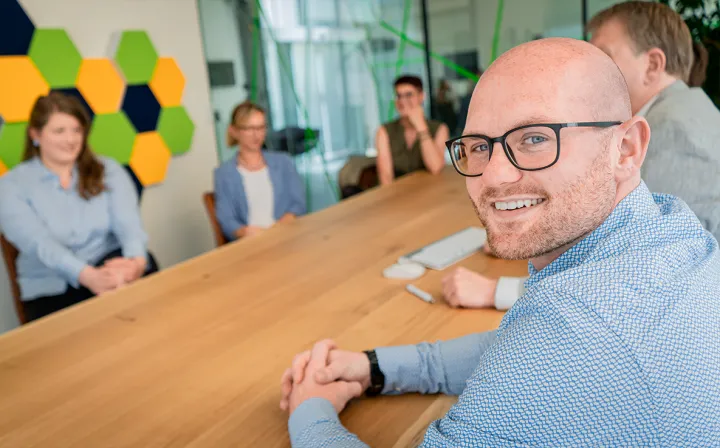 Ein Mann mit Brille schaut in einer Besprechung über die Schulter in die Kamera; im Hintergrund vier Kolleginnen und Kollegen am Konferenztisch.